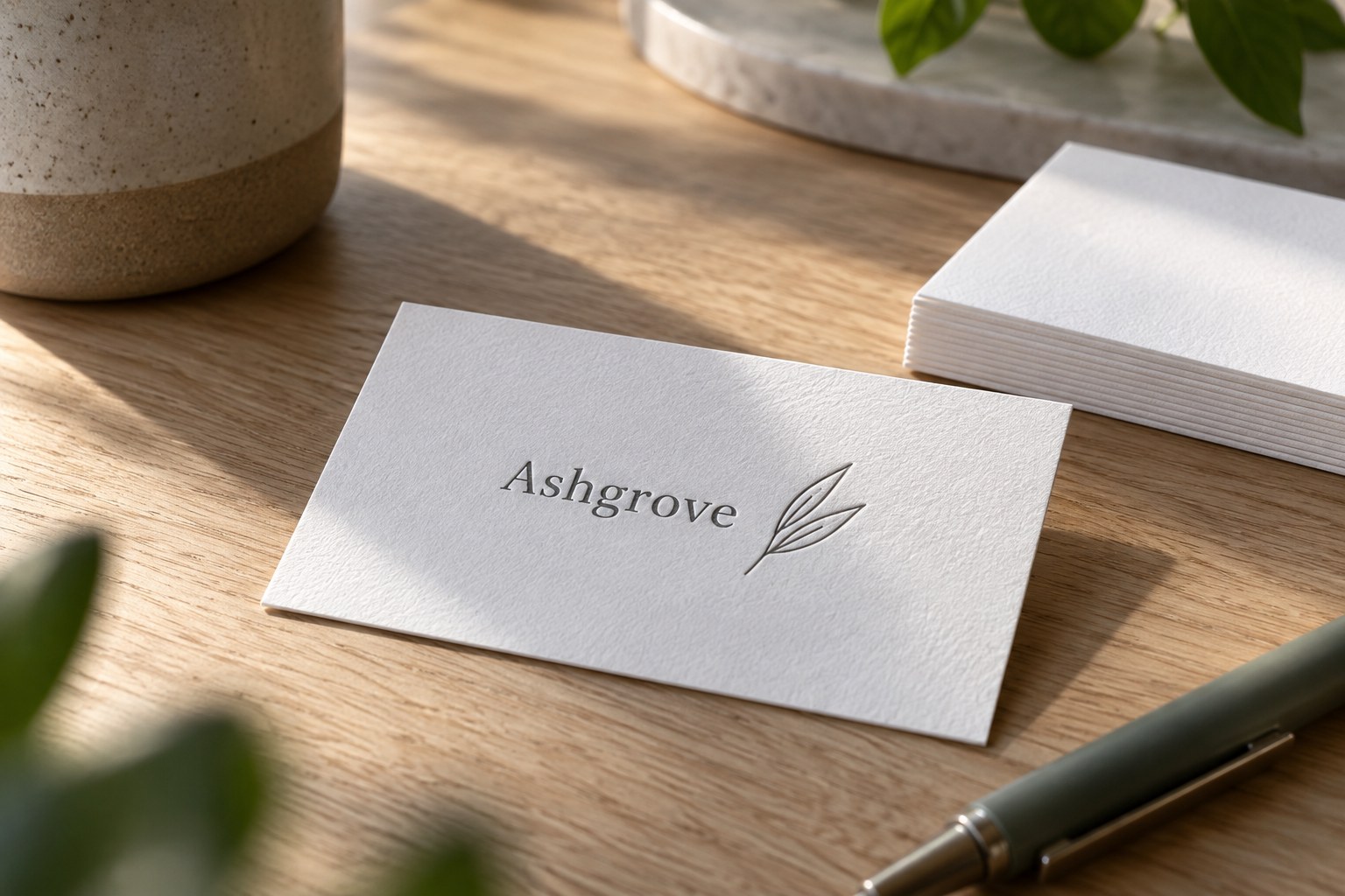 White cotton business card with charcoal pressed lettering and a leaf line drawing on a pale oak desk beside blank white cards and blurred leaves.