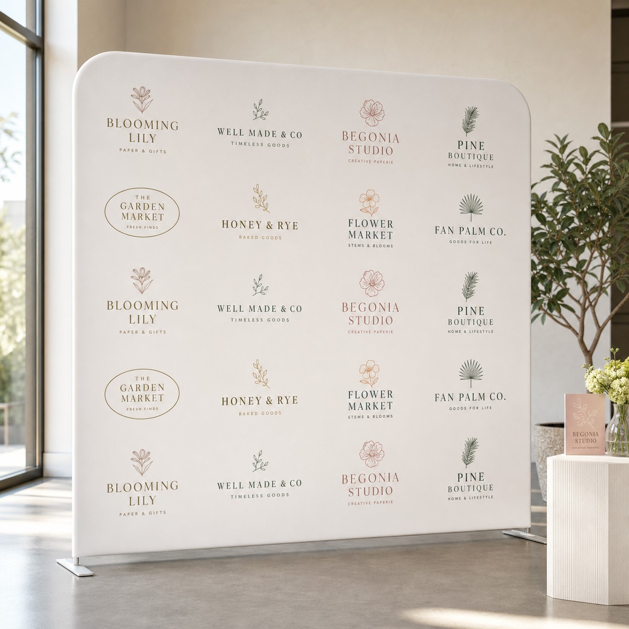 Freestanding stretch fabric media wall printed with a grid of small logos in muted green, pink and brown beside a tall window.