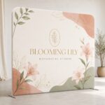 Freestanding stretch fabric media wall with rounded top corners and flat metal feet, printed with peach and sage florals and gold botanical lettering.