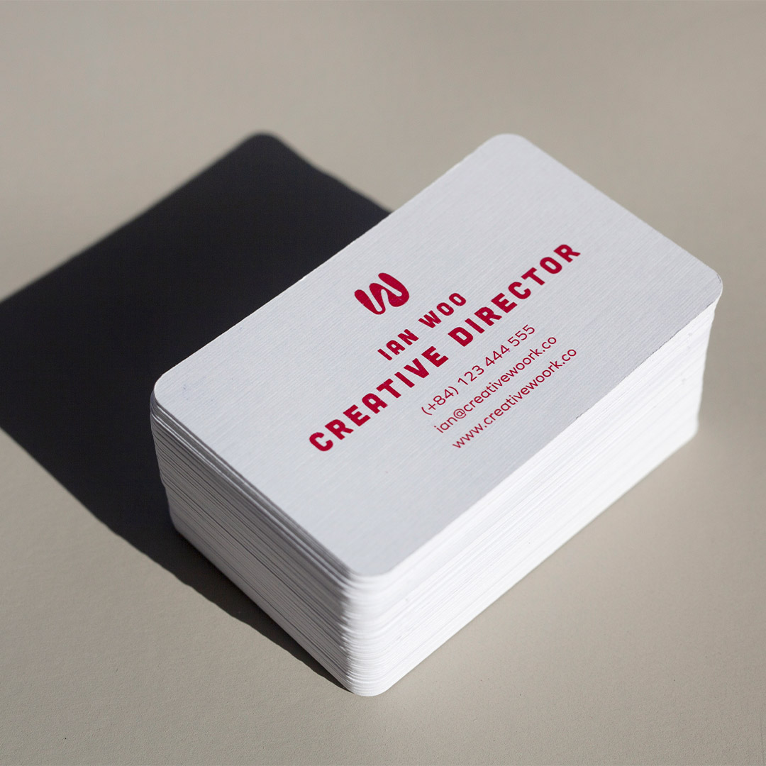 A neat stack of white business cards with rounded corners, a red logo, a name and contact lines on a gray surface with a dark sheet behind.