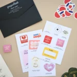 White sample sheets hold stickers and labels beside a black Paperlust Print Shop envelope and loose stickers on a beige surface.