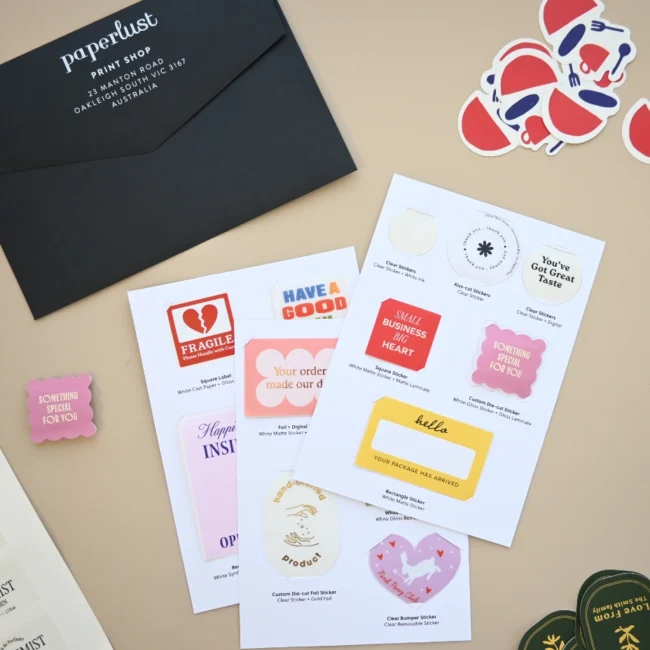 White sample sheets hold stickers and labels beside a black Paperlust Print Shop envelope and loose stickers on a beige surface.