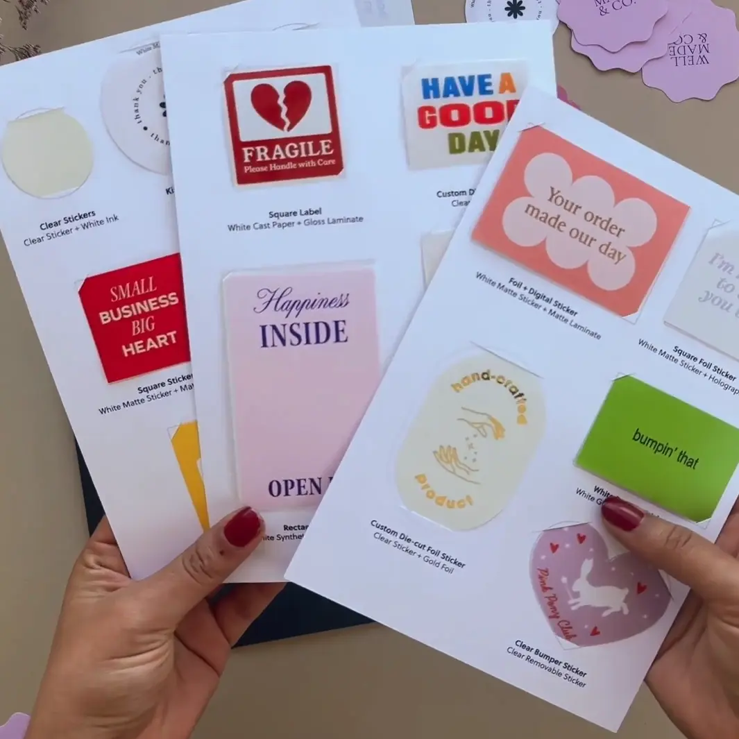 Two hands hold three overlapping white sample sheets showing a red fragile label, a pink rounded label and a heart-shaped sticker.