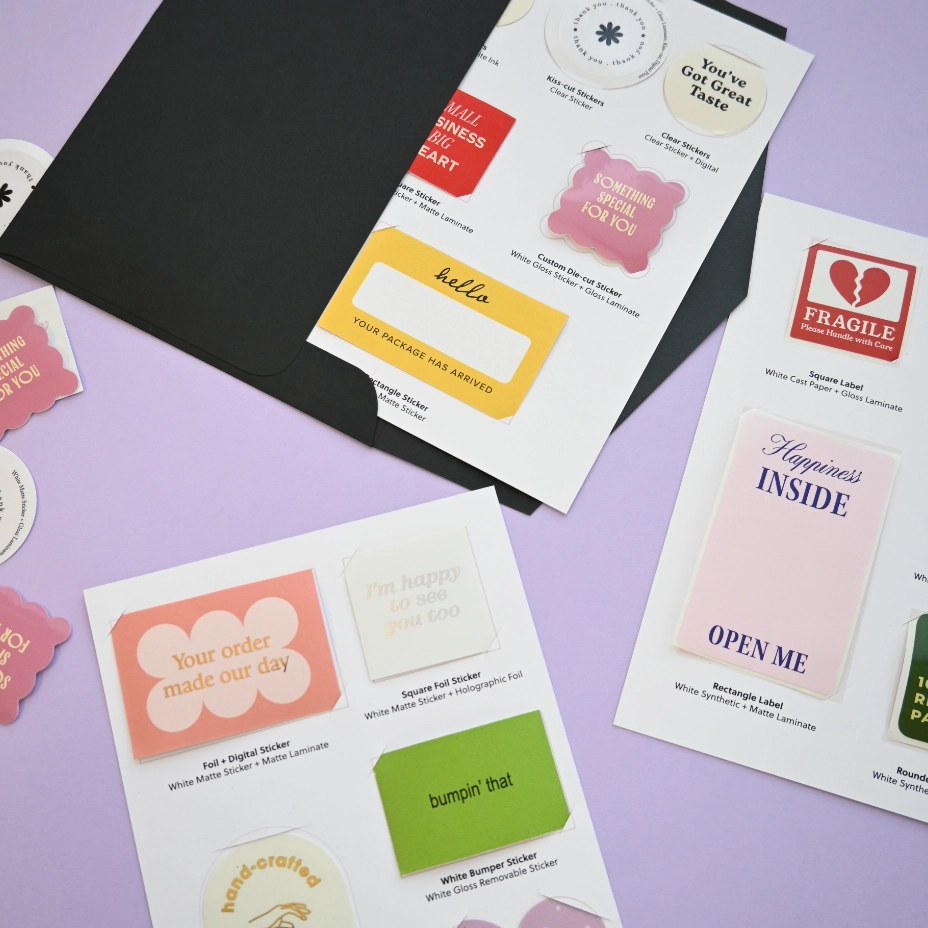 White sample sheets on a lilac surface beside a black envelope display sticker and label samples with printed descriptions underneath.