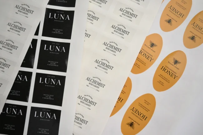 Overlapping sticker sheets: black labels with white lettering, white labels with dark lettering, and orange oval labels with a bee illustration.