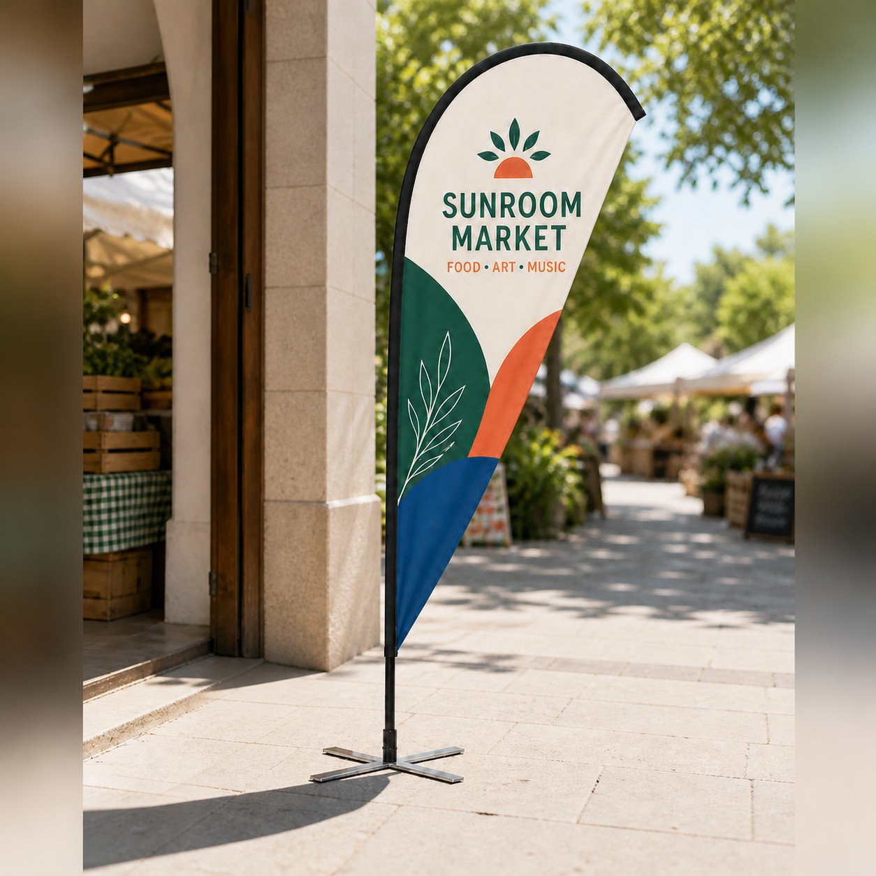 Teardrop banner with white, teal, orange and navy print on a black pole with cross base at an outdoor market on paving with trees.