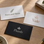 Three cards on a pale timber table: white with sage fern, white with rose gold foil, and black with silver foil, beside a green pen and leaves.