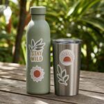 A sage green water bottle with two white-bordered stickers and a stainless steel tumbler sit on a weathered wooden table with blurred tropical garden behind.