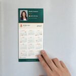 2027 DL fridge magnet calendar in Real estate agent layout on a white fridge, hand pressing it flat, orange holiday dots