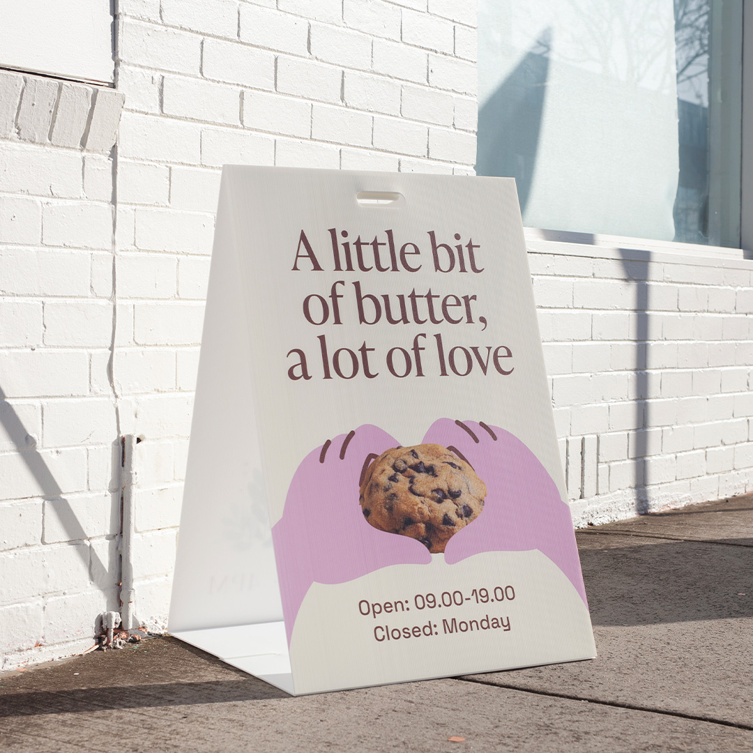 Frameless white corflute A-frame with a cut-out handle stands on a footpath against a white brick wall, printed with bakery lettering and a cookie.