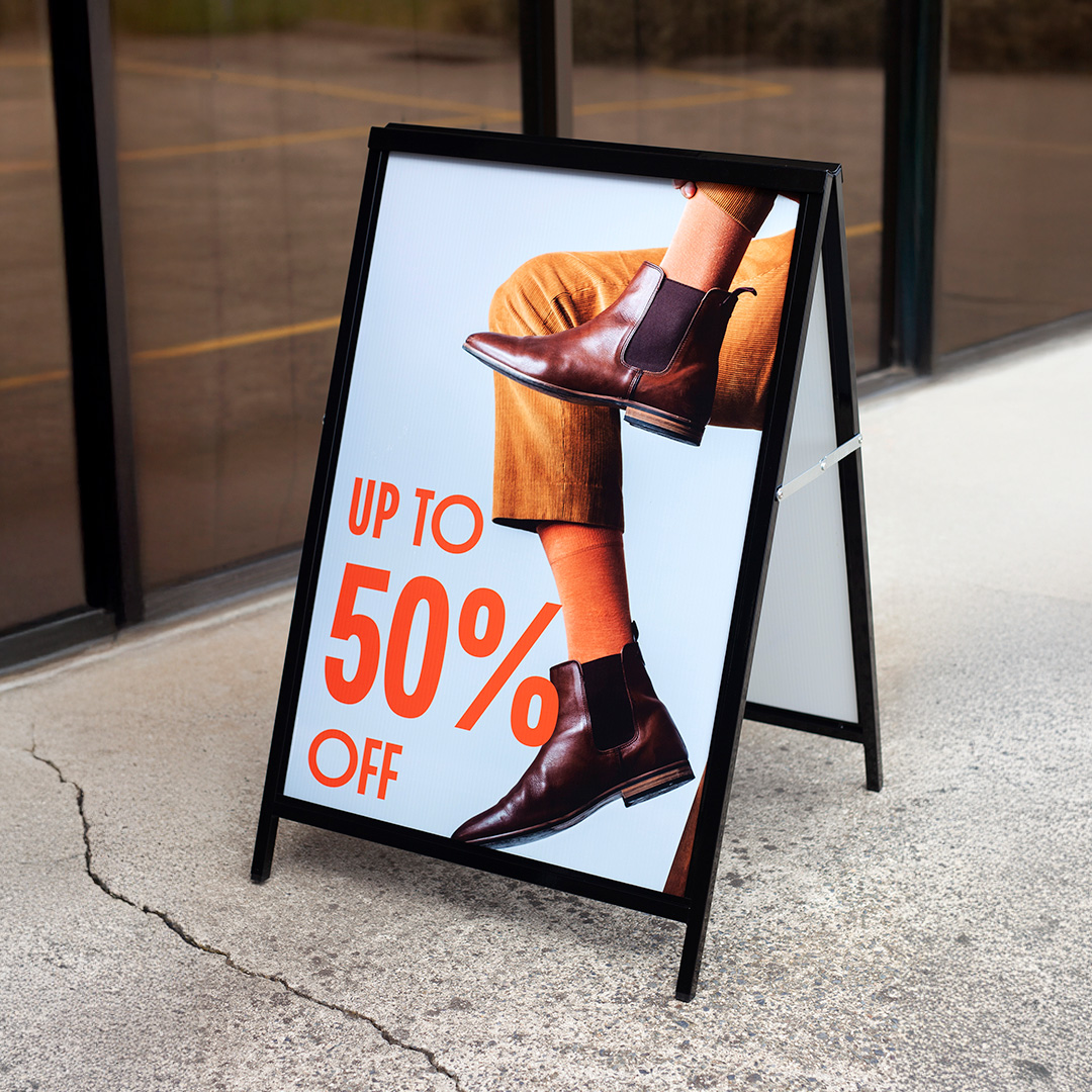 A black steel A-frame with a retail sale panel stands on cracked concrete outside a shop window, the glass reflecting the street.