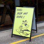 A black steel A-frame with a lime yellow cafe panel stands open on a concrete footpath outside a glass shopfront, another folded A-frame behind it.