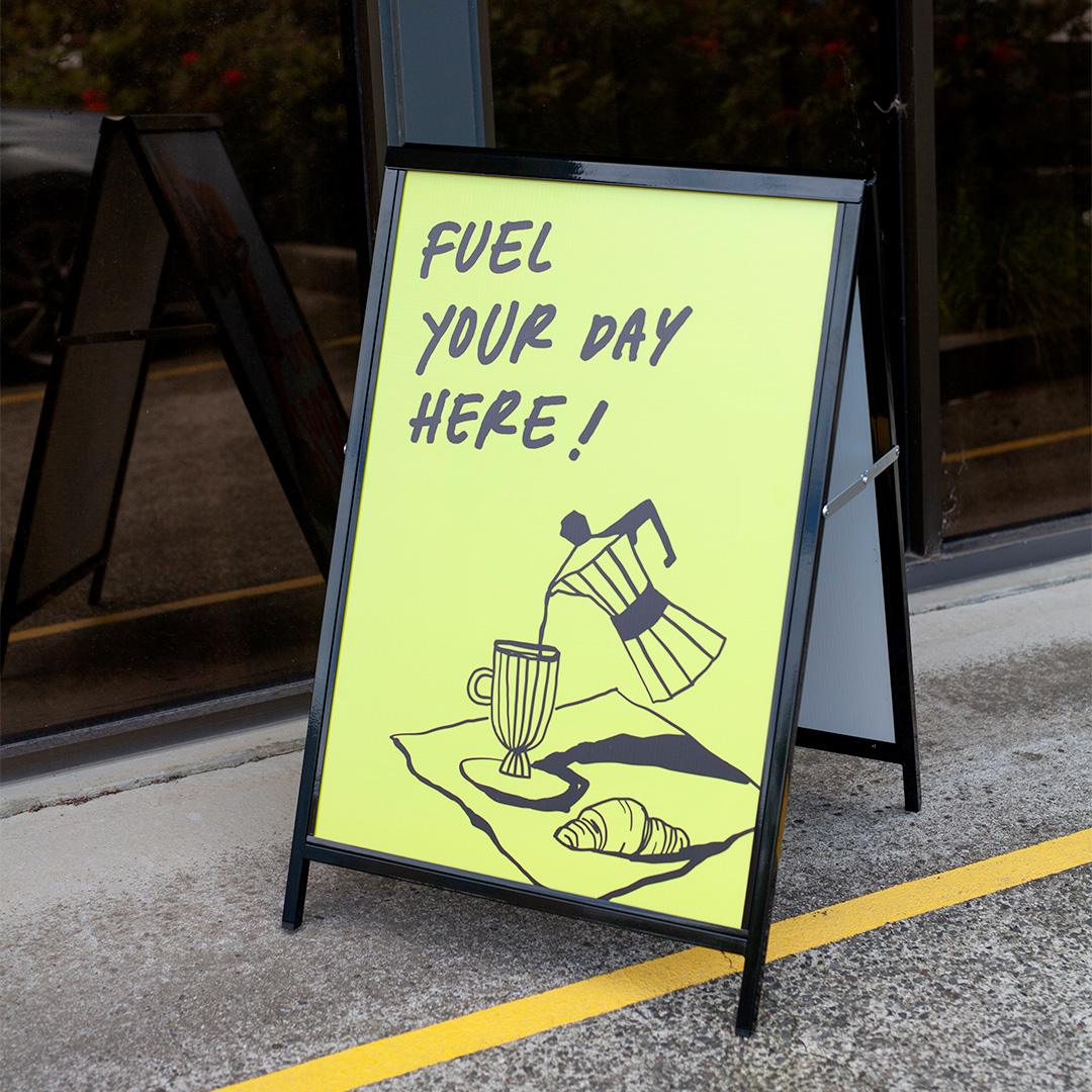 A black steel A-frame with a lime yellow cafe panel stands open on a concrete footpath outside a glass shopfront, another folded A-frame behind it.
