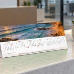 Closed A4 2027 tent desk calendar with coastal photo above first six months on stone counter beside succulent