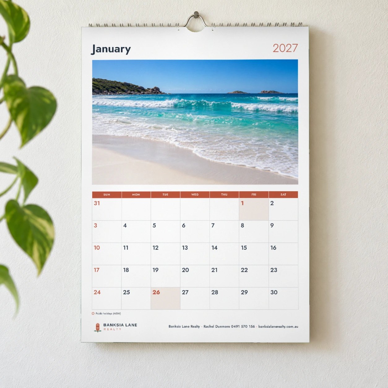 A4 2027 wall calendar on white wall, turquoise beach photo under January and 2027, red day name row, logo and details footer