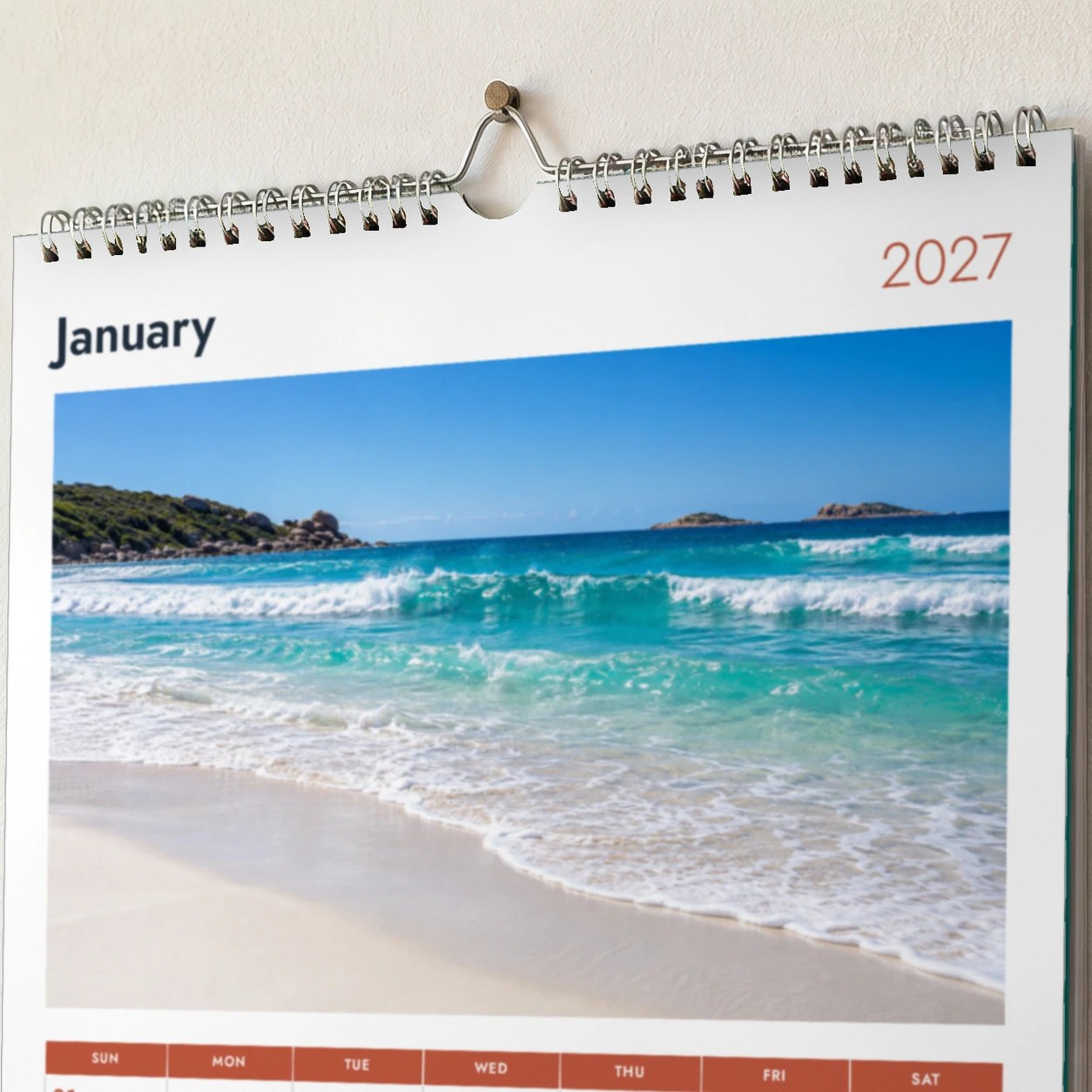 Close detail of A4 wall calendar top, silver twin loop wire, wire hanger on nail, January heading and 2027 over beach photo