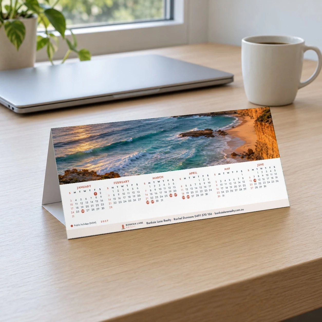 A4 2027 tent desk calendar showing a coastal photo above January to June on timber desk beside laptop and coffee mug