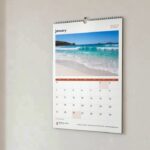 A4 wall calendar in Classic layout on pale hallway wall, beach photo above January grid, dried grass in a vase beside it