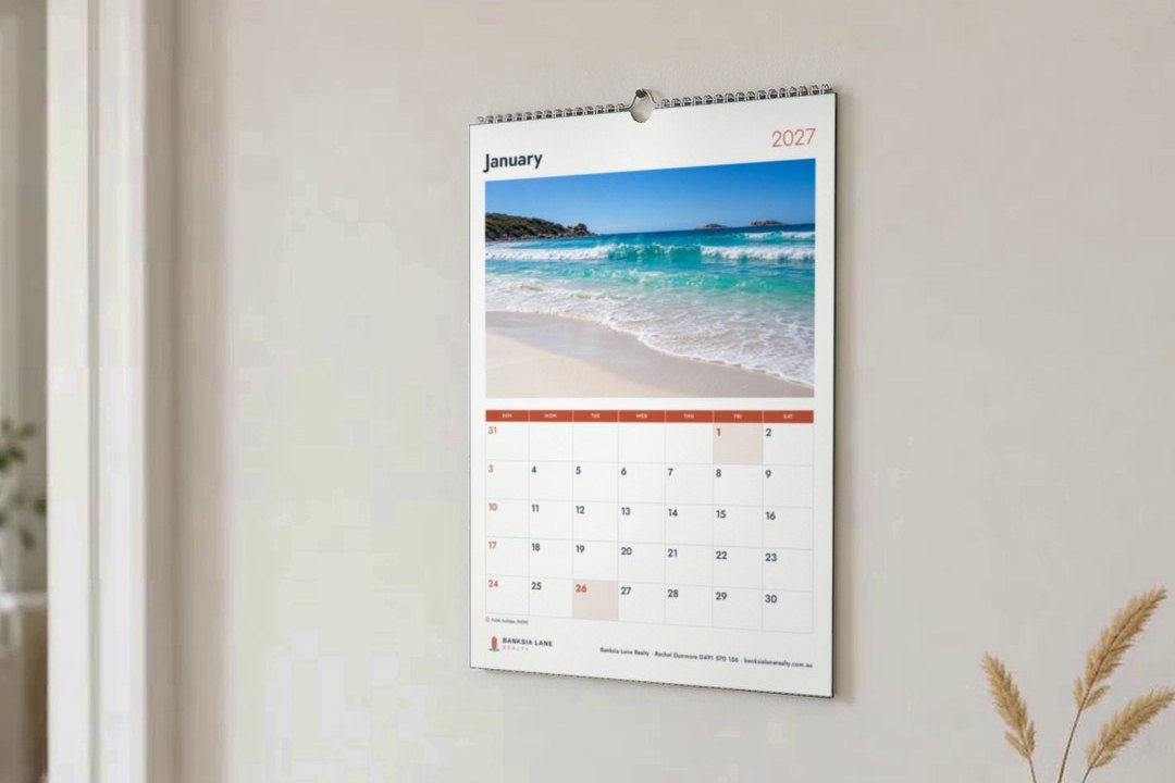 A4 wall calendar in Classic layout on pale hallway wall, beach photo above January grid, dried grass in a vase beside it