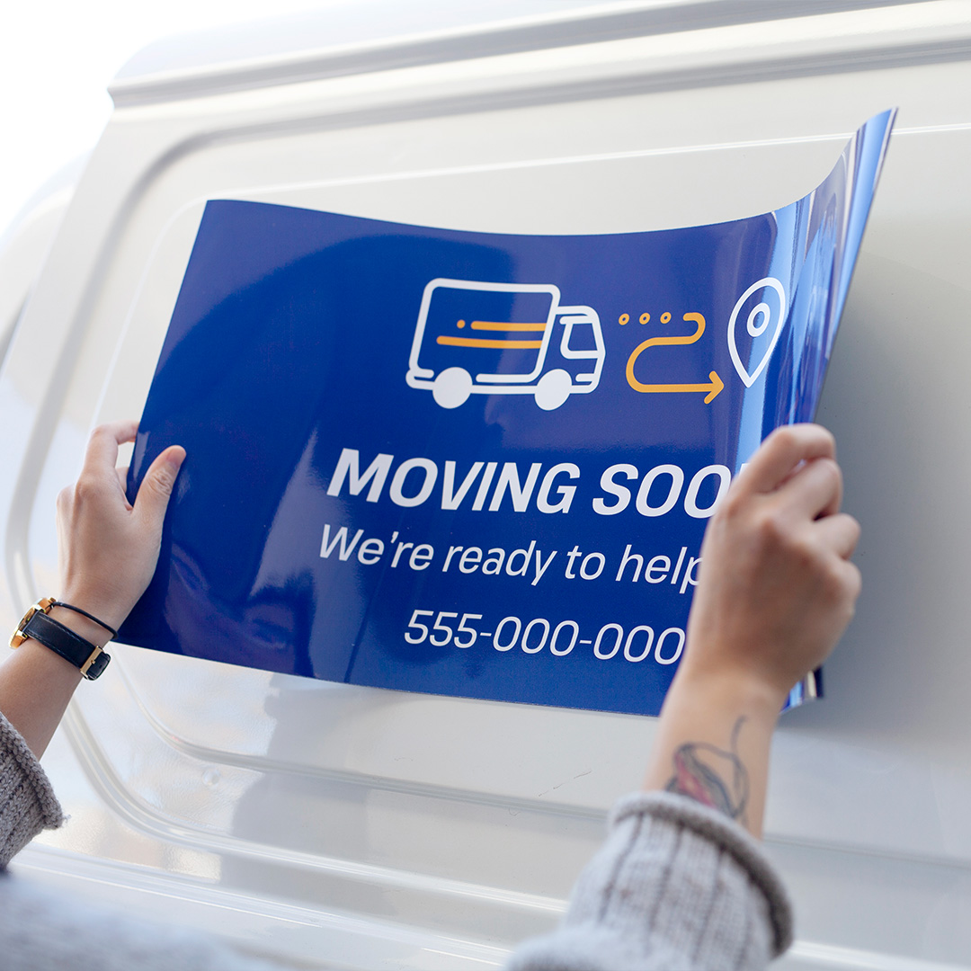 Hands placing a royal blue printed magnetic sign on the white side panel of a van, with one corner still lifting away.