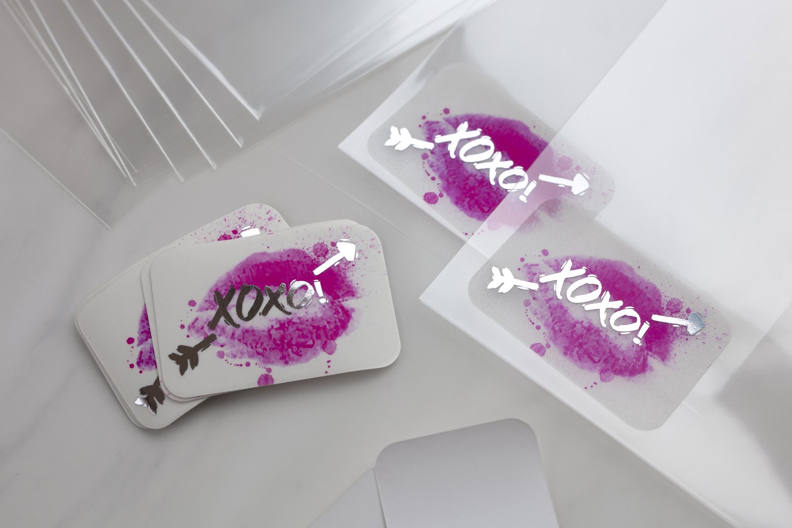 On a white marble surface, opaque white stickers with a magenta lipstick kiss and silver foil lettering sit beside two clear stickers on clear backing, their see-through pink print letting the pale marble show.