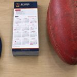 Stack of DL fridge magnet calendars with navy header, key dates box on timber table, red football, navy lanyard whistle