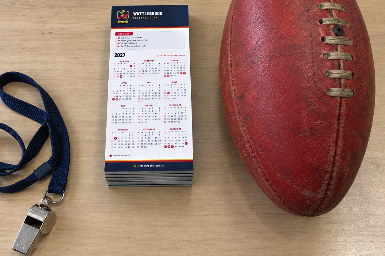 Stack of DL fridge magnet calendars with navy header, key dates box on timber table, red football, navy lanyard whistle