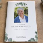 Closed A5 funeral order of service booklet in the Eucalyptus design on a pale oak table with eucalyptus leaves and a rectangular portrait of a smiling older woman