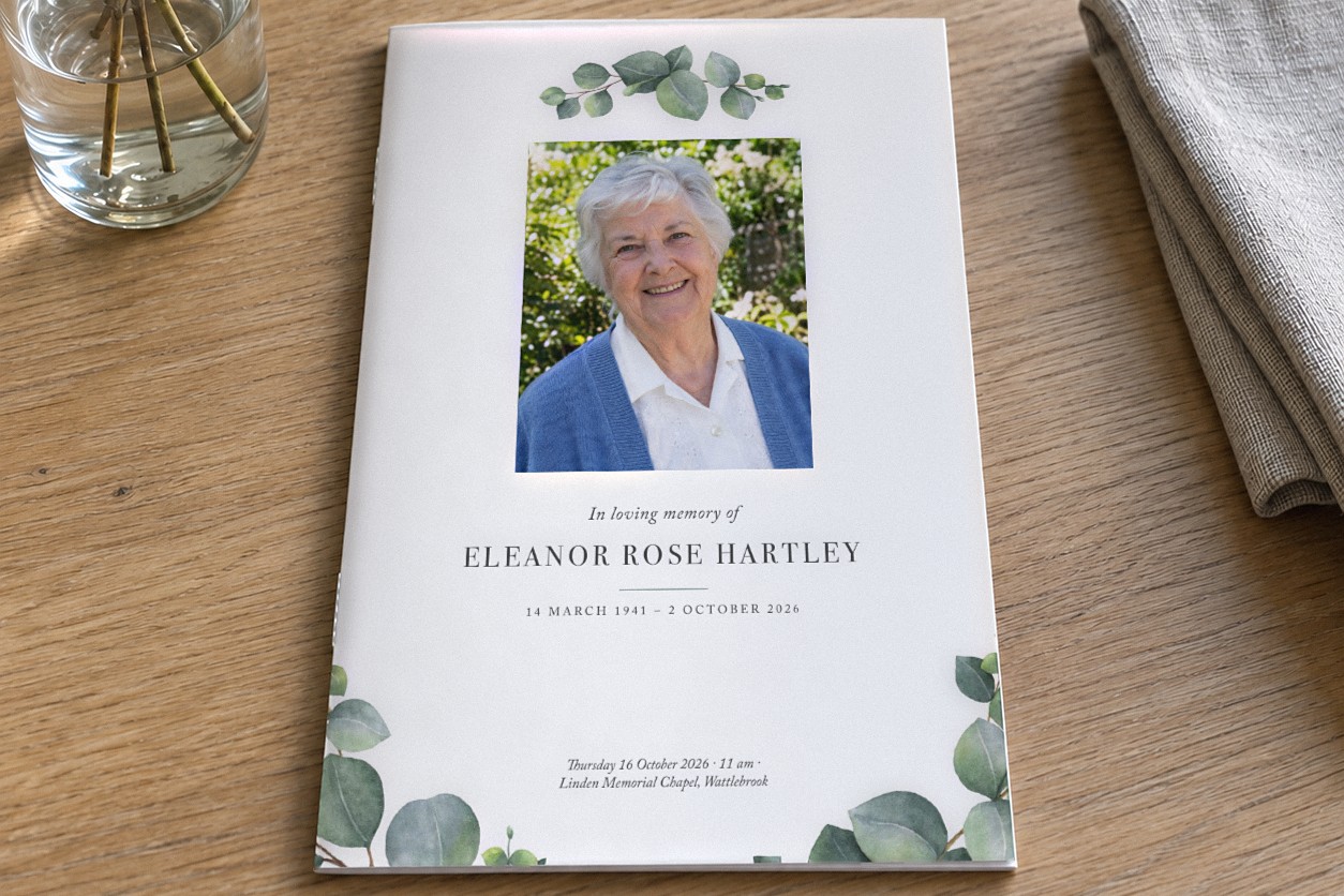 Closed A5 funeral order of service booklet in the Eucalyptus design on a pale oak table with eucalyptus leaves and a rectangular portrait of a smiling older woman