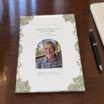 Closed A5 funeral order of service booklet in the Visco Garden design on a dark polished timber table with olive green botanical flowers framing an arch shaped portrait of a smiling older man