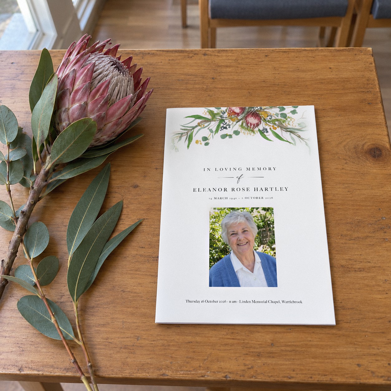 Closed A5 funeral booklet in the Sarulean design on a warm timber table with a garland of Australian natives above a rectangular portrait of an older woman