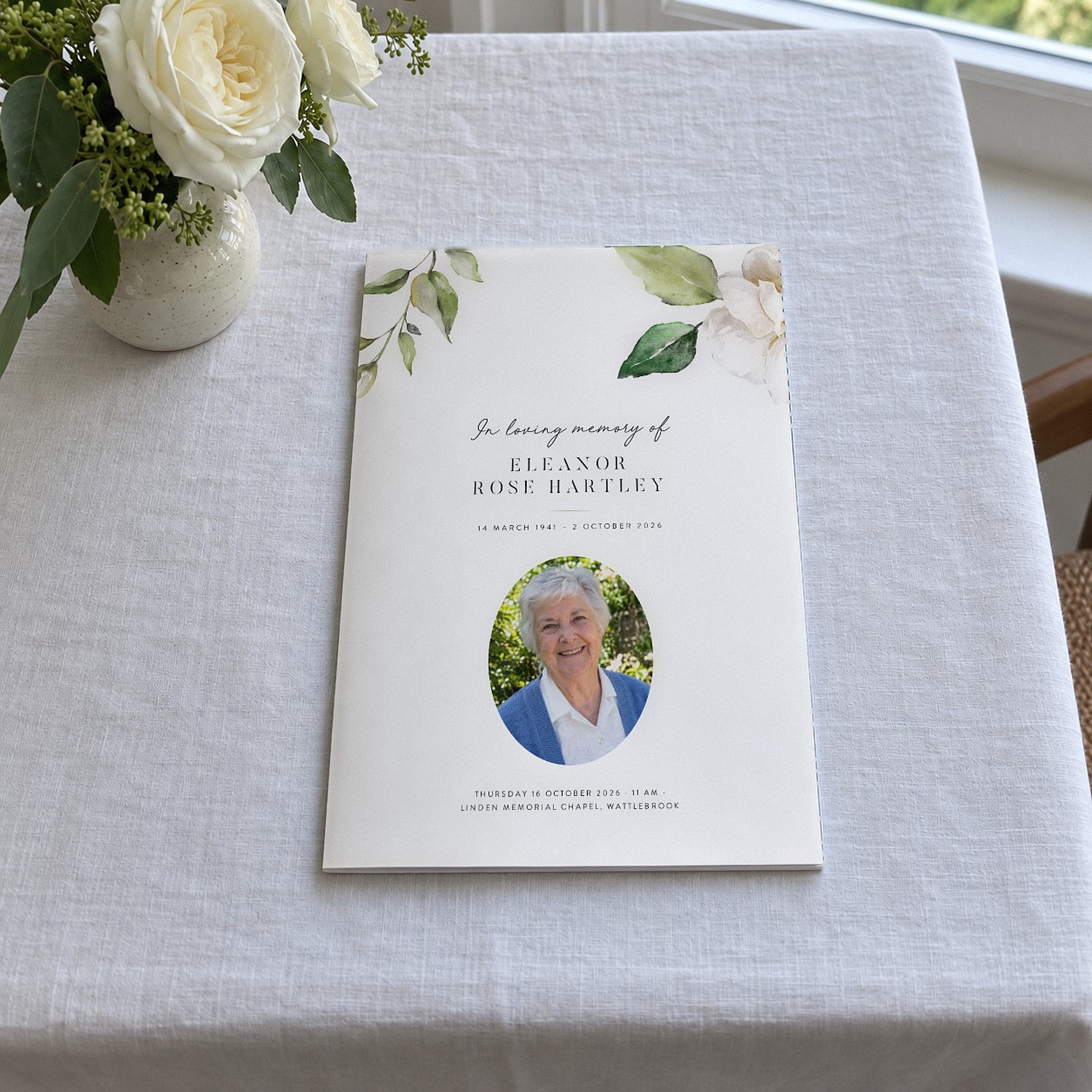 A closed A5 booklet sits on white linen, with a watercolour white rose and green leaves framing an oval portrait of an older woman