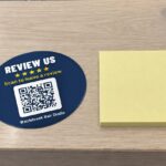 Review QR code sticker on a timber counter with REVIEW US, five gold stars and a QR code by a yellow sticky note pad
