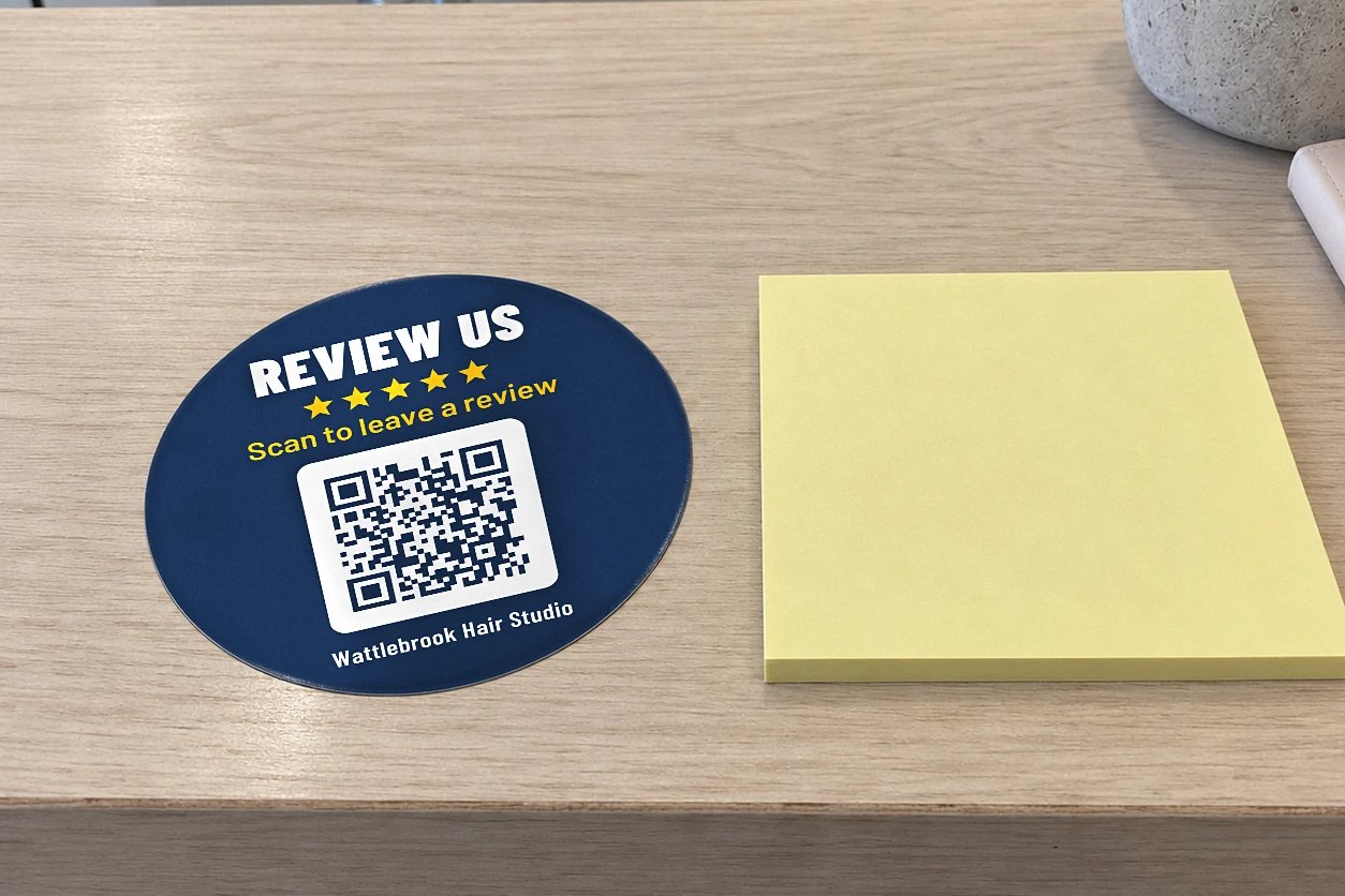 Review QR code sticker on a timber counter with REVIEW US, five gold stars and a QR code by a yellow sticky note pad