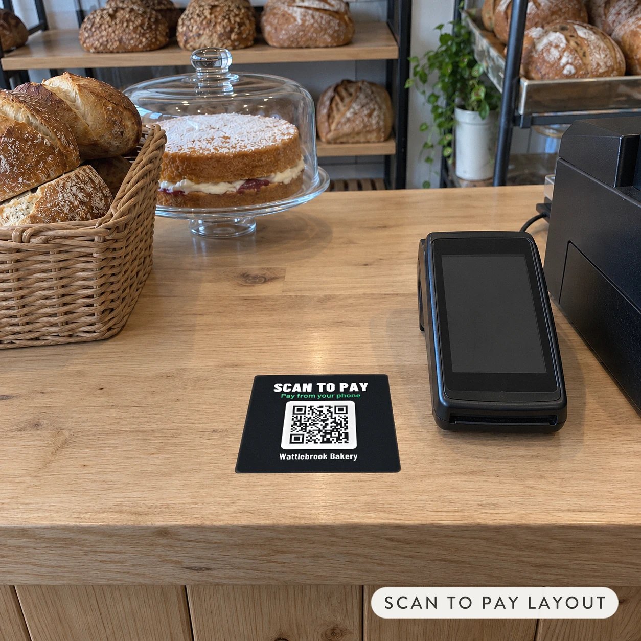 A black square Scan to pay QR code sticker on a counter beside a bakery payment terminal