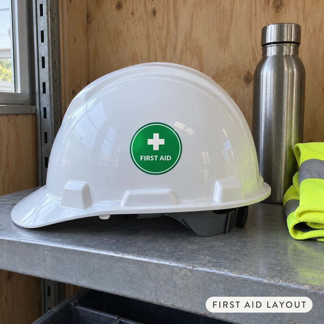 A green round first aid hard hat sticker with a white cross on a safety cap beside a drink bottle