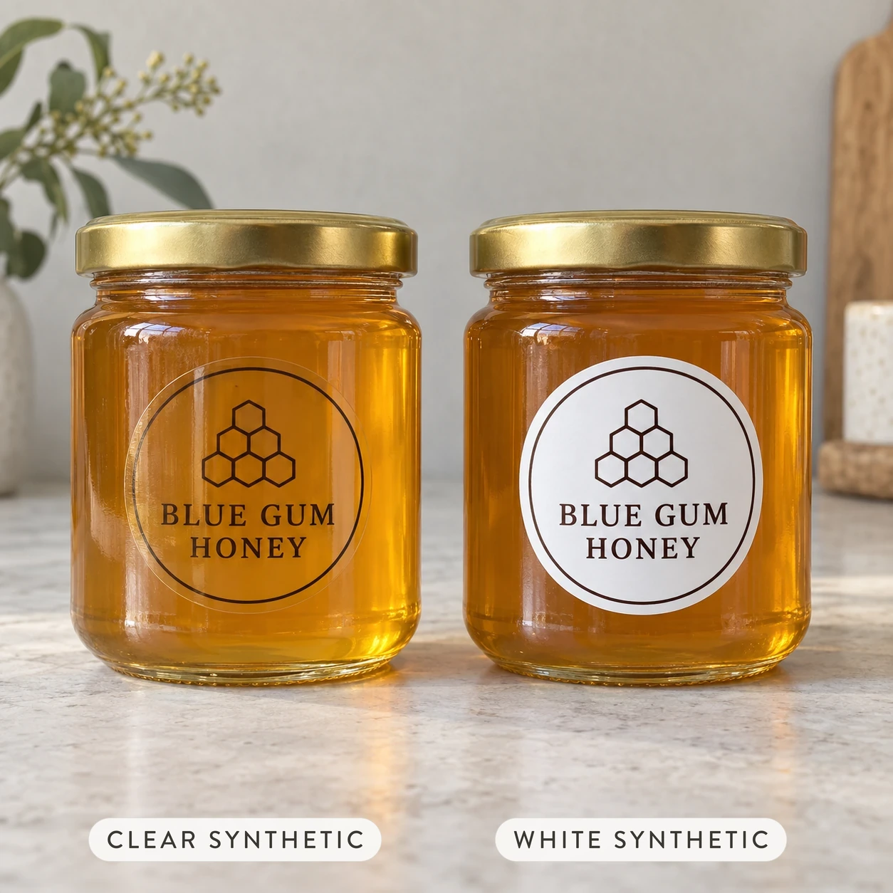 Two golden honey jars with gold screw lids on a pale stone bench, one clear circle label and one white circle label with matching honeycomb icon