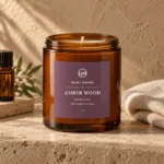Amber glass candle jar with visible wick on a pale stone ledge, plum purple rectangle label with white text, oatmeal cloth and green leaves behind