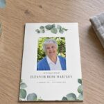 Memorial card with watercolour eucalyptus sprays, a smiling older woman photo and In loving memory of on a pale oak table