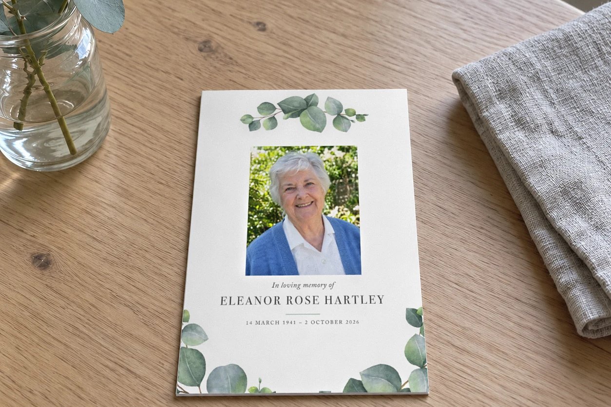 Memorial card with watercolour eucalyptus sprays, a smiling older woman photo and In loving memory of on a pale oak table
