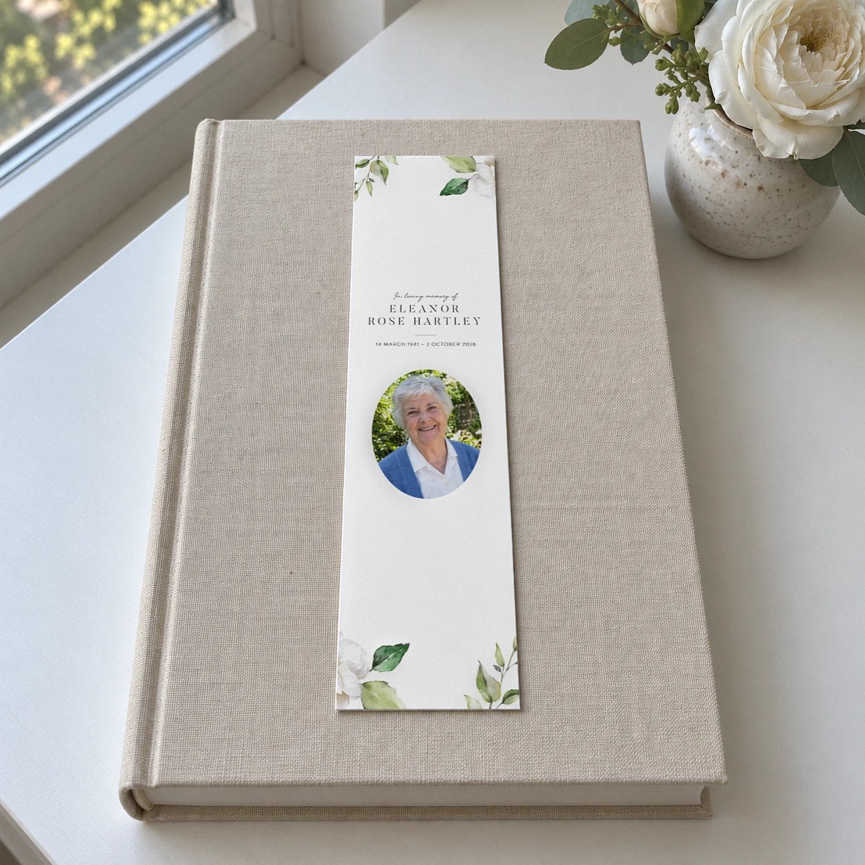 Memorial bookmark with an oval photo of a smiling older woman, white roses and green leaves on a linen covered book