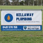 A white and blue vinyl sponsor banner cable tied through eyelets to a galvanised chain wire fence along a suburban oval