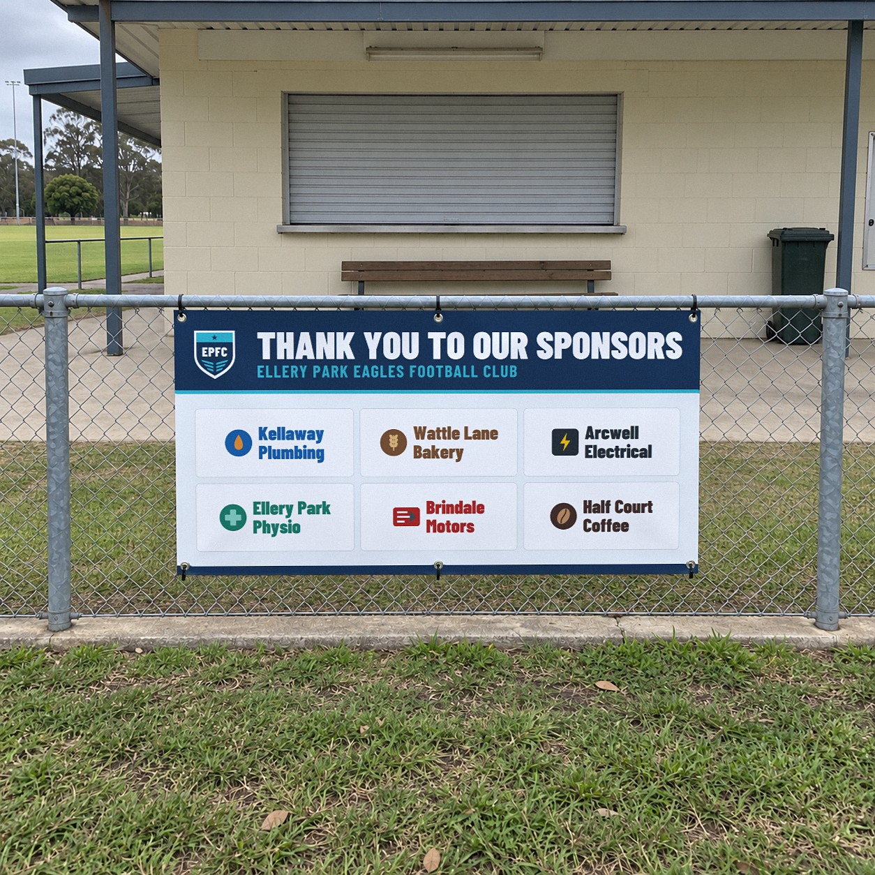 Six sponsor logos in white panels on a vinyl sponsor board fixed to a chain wire fence beside a concrete path