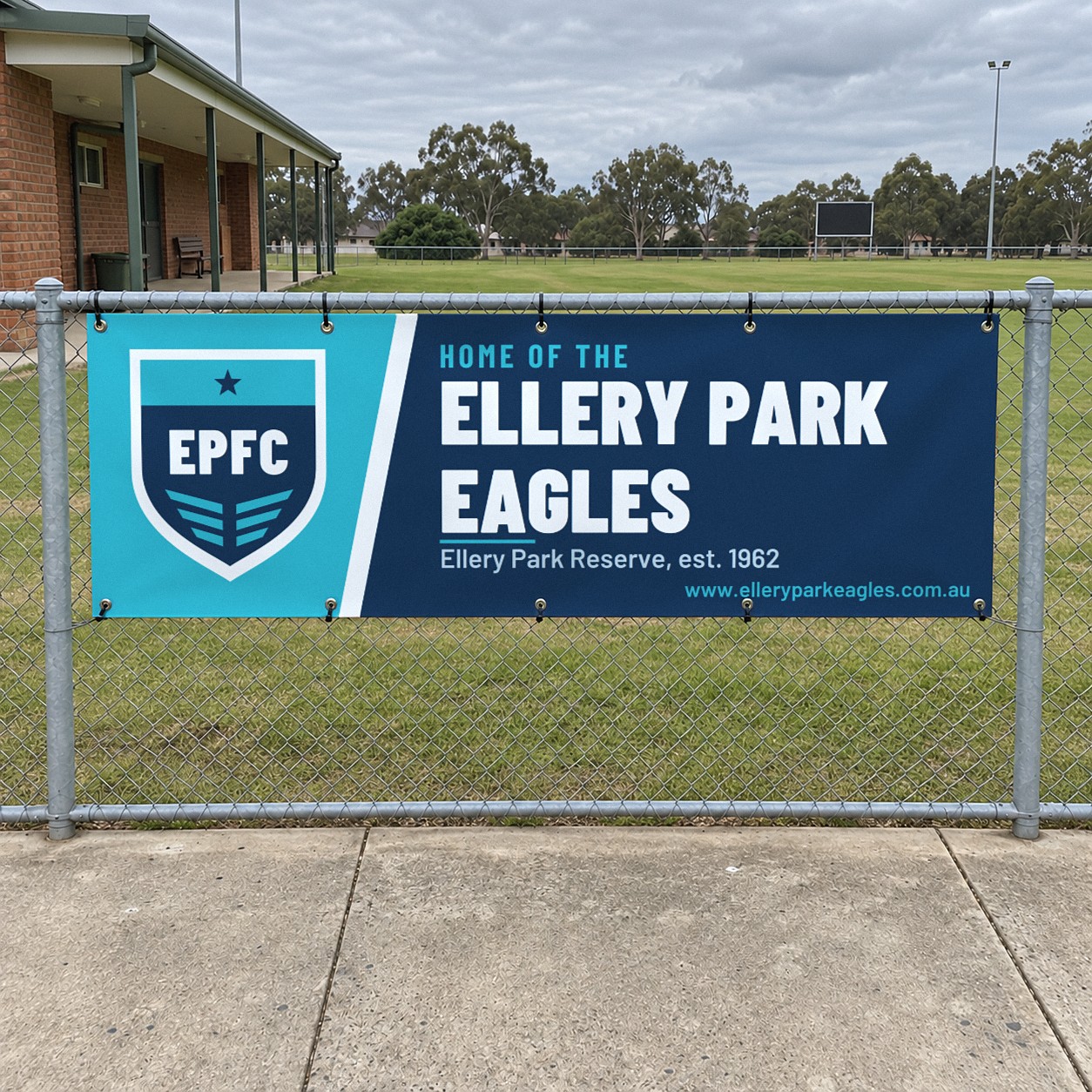 Navy and aqua vinyl club banner with a club crest and white club name on a slanted aqua panel, fixed through eyelets to a chain wire fence near a sports oval