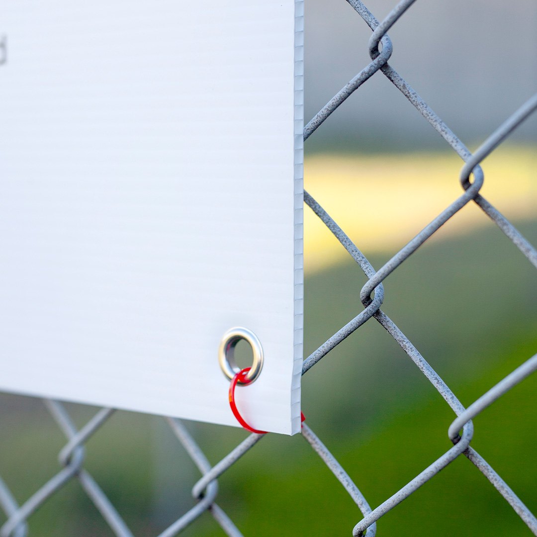 Corflute sign corner with one round silver eyelet and red cable tie on a chain wire mesh fence