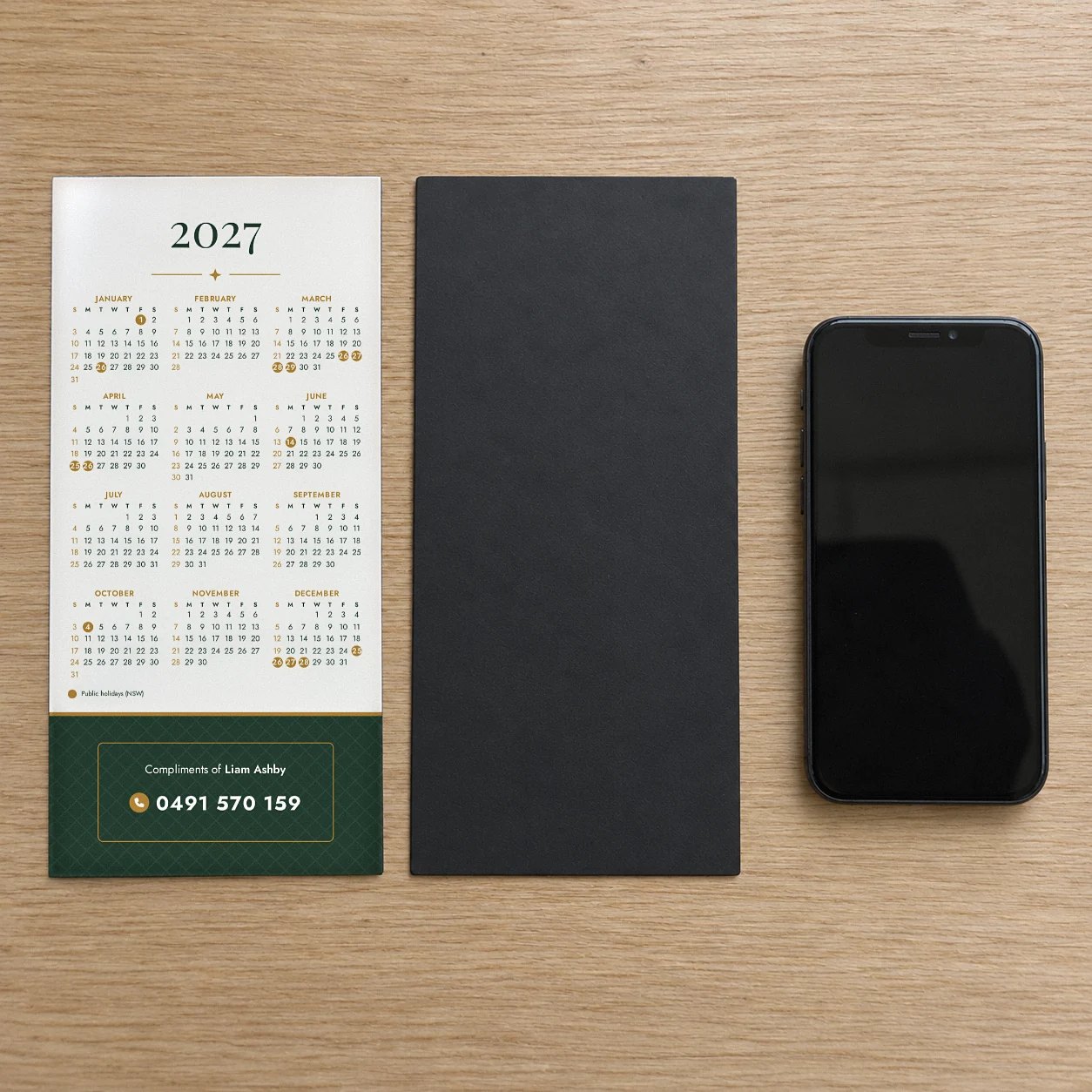 Two DL fridge magnet calendar prints on timber, one face up with 2027, other face down, black magnet back, phone beside
