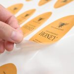Fingers lift the pointed end of an amber oval label from a white backing sheet, the curled edge showing its pale underside, more bee labels blurred behind.