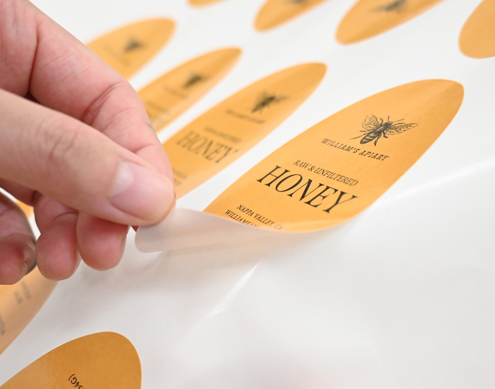 Fingers lift the pointed end of an amber oval label from a white backing sheet, the curled edge showing its pale underside, more bee labels blurred behind.