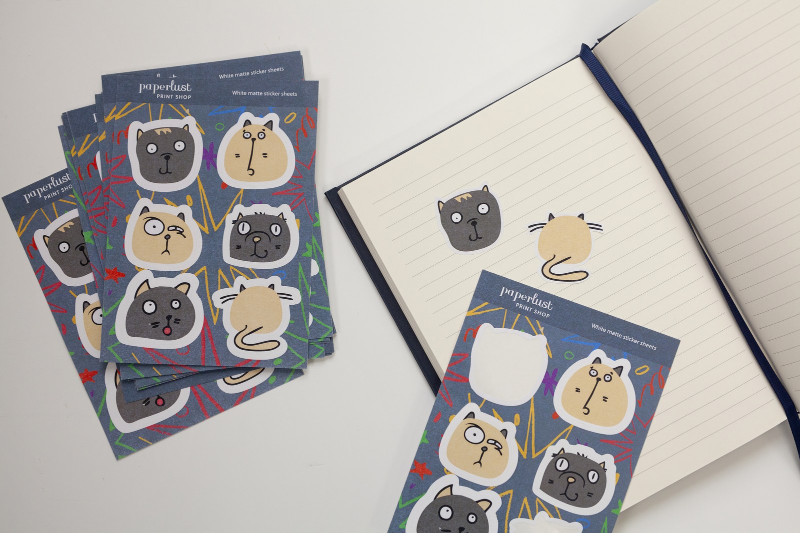Cartoon cat face sticker sheets fanned on a white desk · Sticker Sheets in White Matte Sticker stock · stickers kiss cut through the top layer